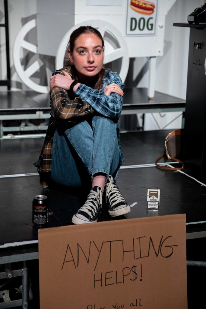 Student girl sitting down with a drink can, a sign that say "Anything Helps" and empty packet of cigarettes acting to be homeless