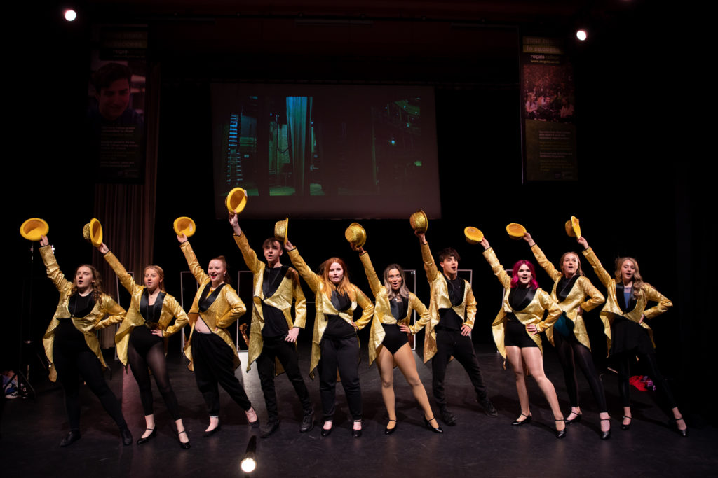 Students in a line, wearing gold costumes, lifting their gold hats up with their hands.