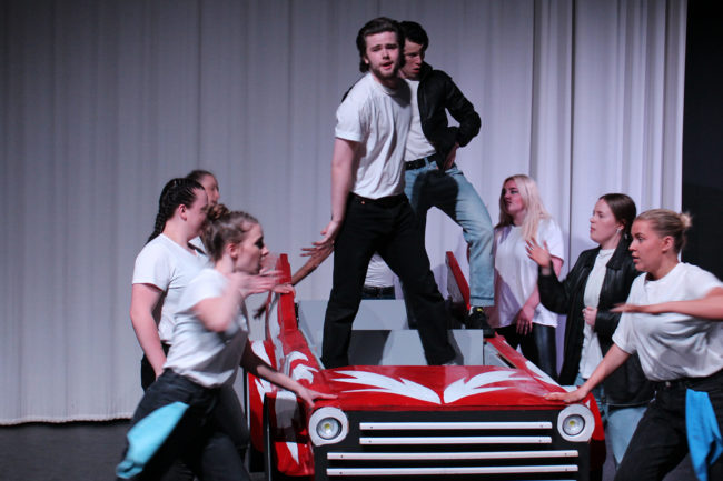 Grease performance 2022 - students dancing around a prop stage car, two students standing in the stage care