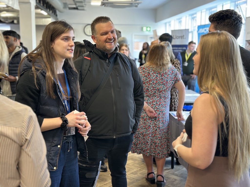 An employer speaking to students and their parents on their stand at the Meet the Employer event. Many people can bee seen in the background.